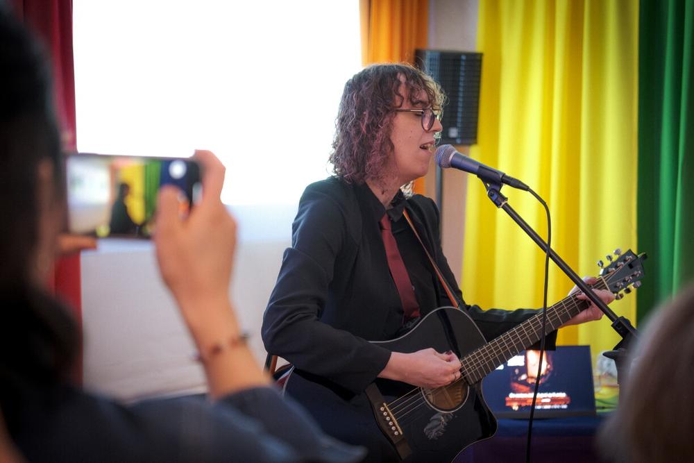 Mia Vale, performing as Cherry Red, singing with a guitar in front of rainbow curtains while a guest films on a phone